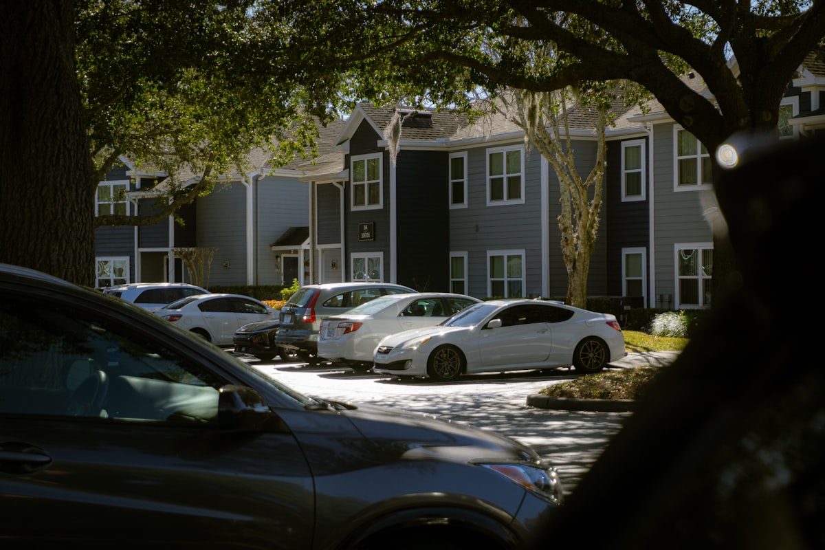 Rows of cars parked in an apartment complex parking lot under a shade tree, with no private driveways in sight