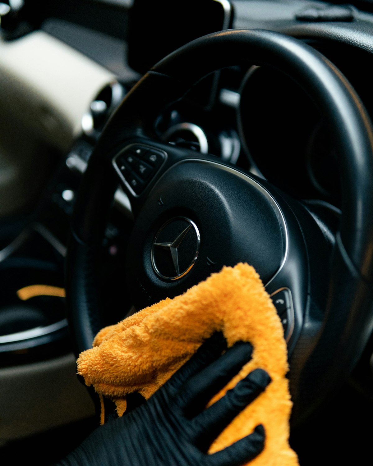 A gloved detailer wiping down a car's steering wheel and dashboard with an orange microfiber cloth.