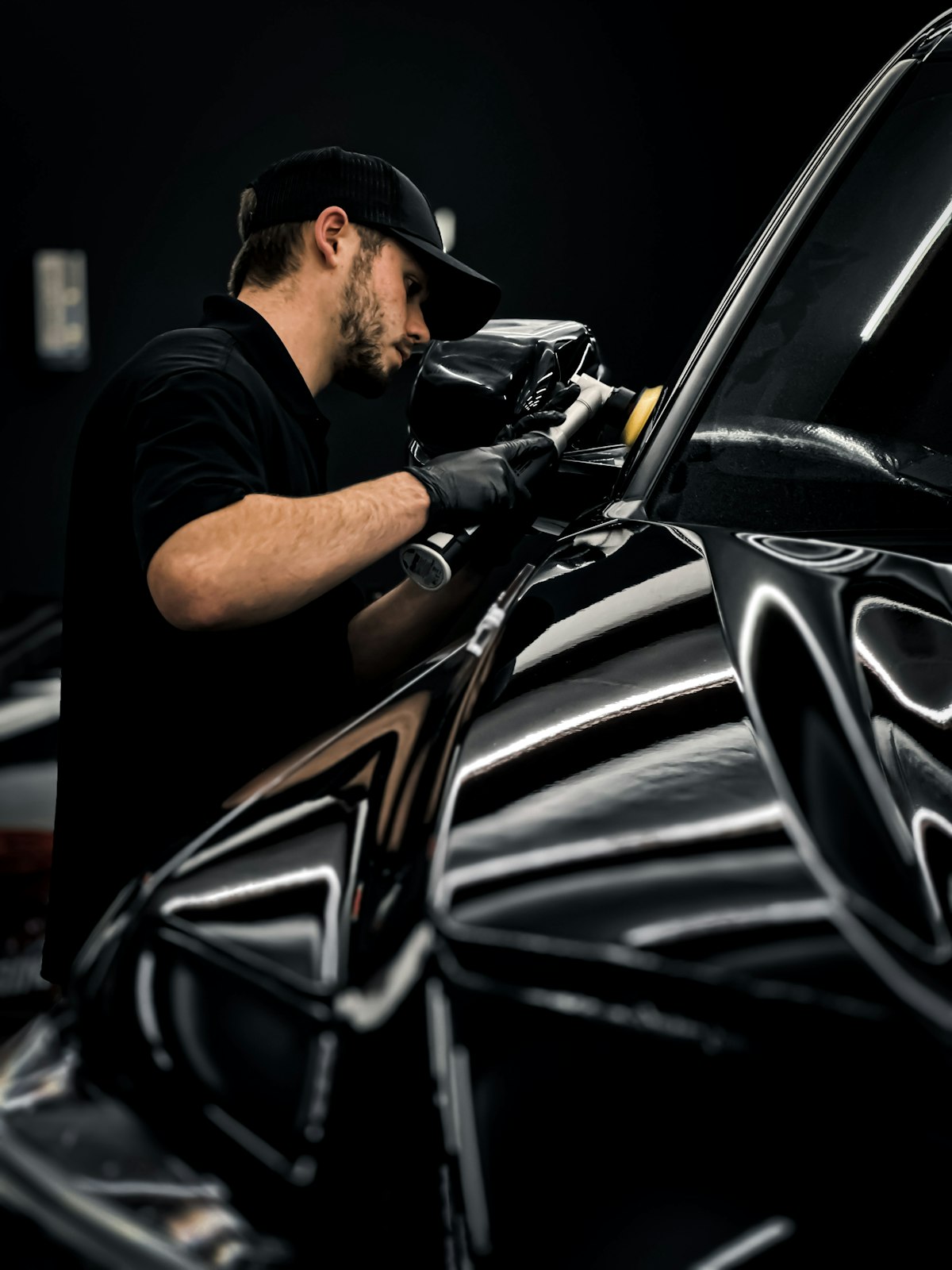 A detailer in a black cap carefully working over the glossy paint of a black car during a full detail.