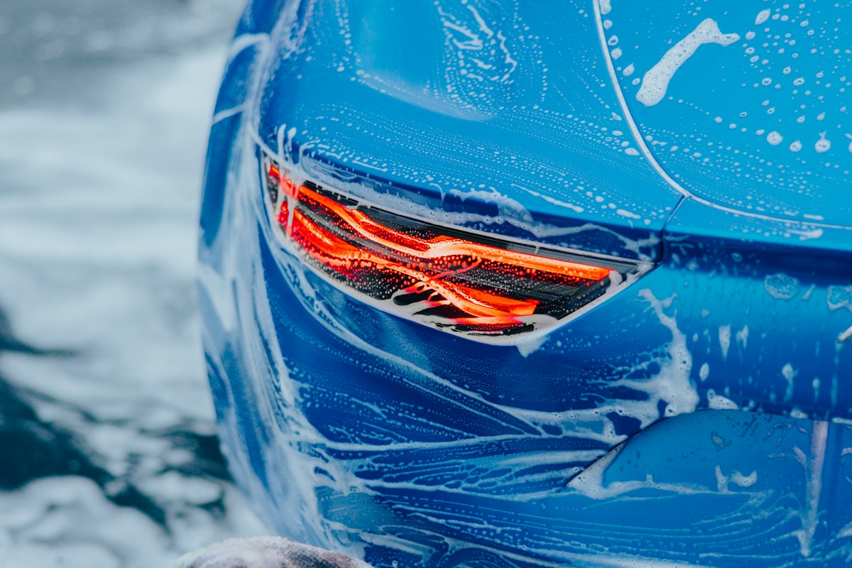 Thick soap foam covering a car's exterior panel and taillight during a hand wash, the first step of a full detail