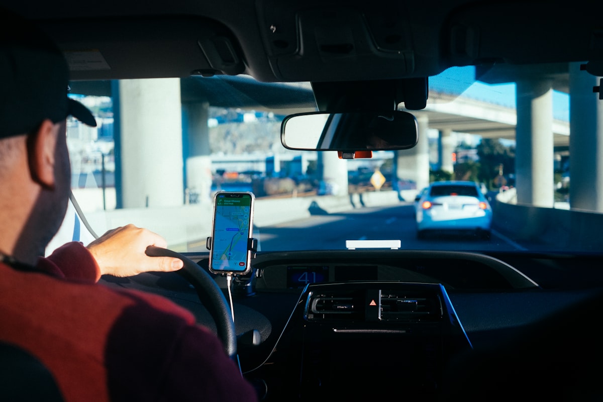 A rideshare driver's hand on the steering wheel with a navigation app mounted on the dashboard, freeway overpass visible through the windshield