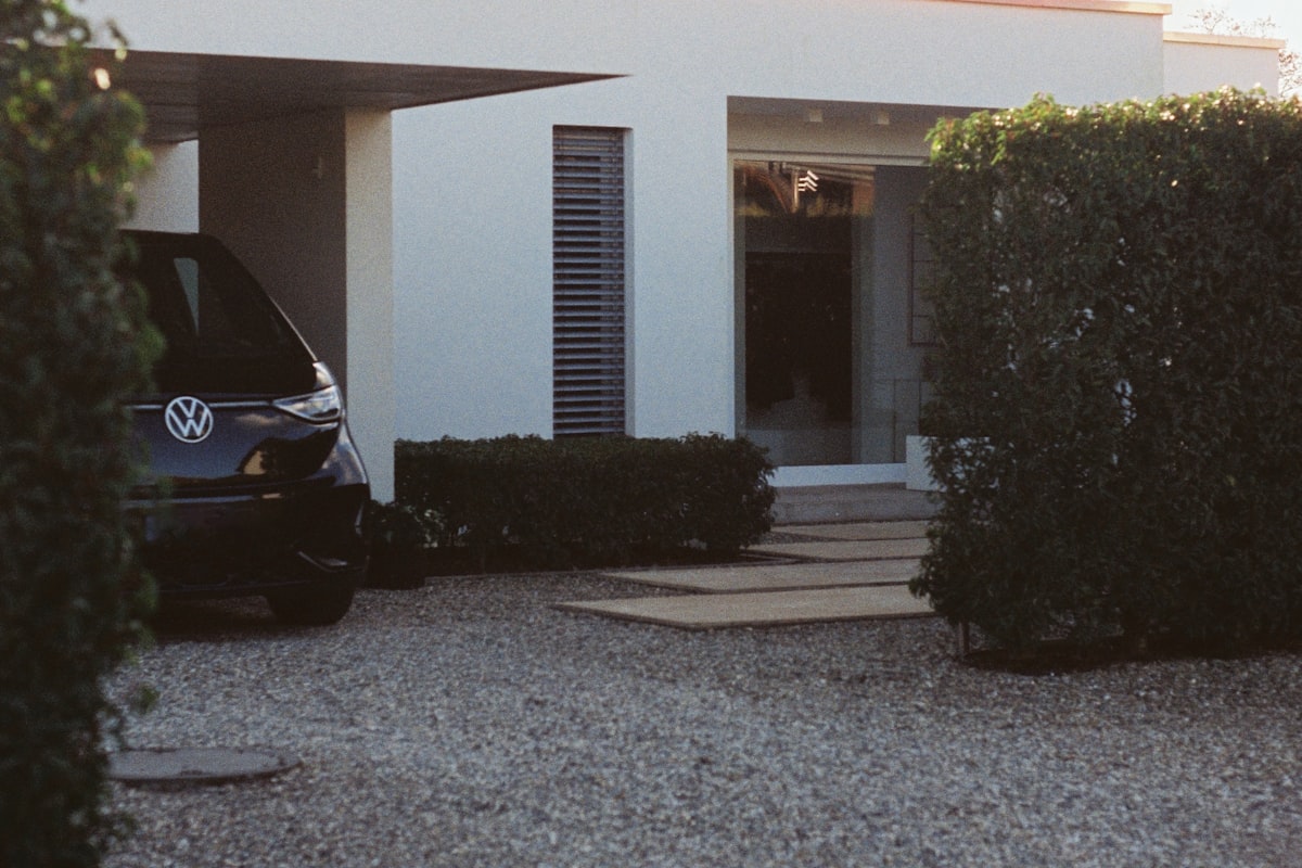 A car parked under a covered carport beside trimmed hedges on a Santa Clara driveway, the kind of landscaped property where sprinkler overspray reaches parked cars