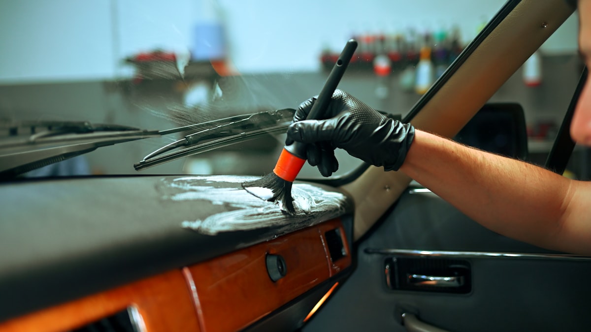 A detailer's gloved hand applying interior cleaner to a sticky car dashboard with a brush